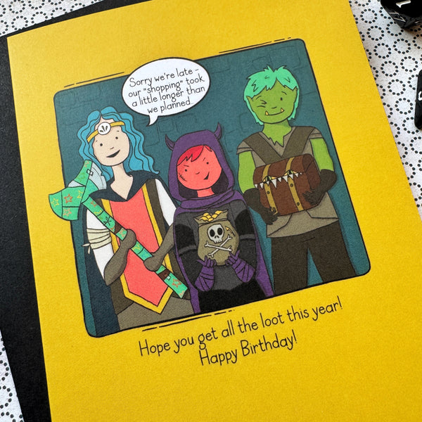 Video shows a closeup look at a yellow greeting card featuring a comic illustration of 3 adventurers - a little beat up, carrying birthday presents that are definitely dungeon loot. One character says "Sorry we're late - our "shopping" took a little longer than we planned." The caption below the illustration reads "Hope you get all the loot this year! Happy birthday!"