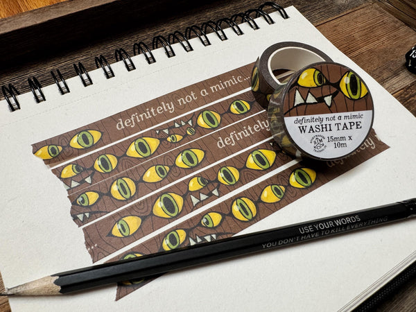 Image of an illustrated comic style washi tape design with a wood background and classic mimic eyes and teeth, and the words &quot;definitely not a mimic&quot;. Also shows 2 packaged rolls and a pencil.