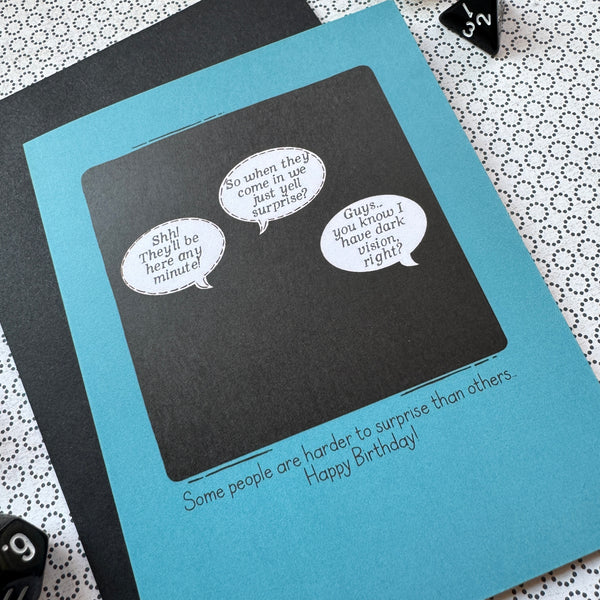 Picture shows a blue greeting card with a comic illustration - a black square with 3 speech bubbles. The first says "Shh! They'll be here any minute!" The second says "So they come in, we just yell surprise?" And the third says "Guys...you know I have dark vision, right?" The caption beneath the illustration reads "Some people are harder to surprise than others... Happy Birthday!"